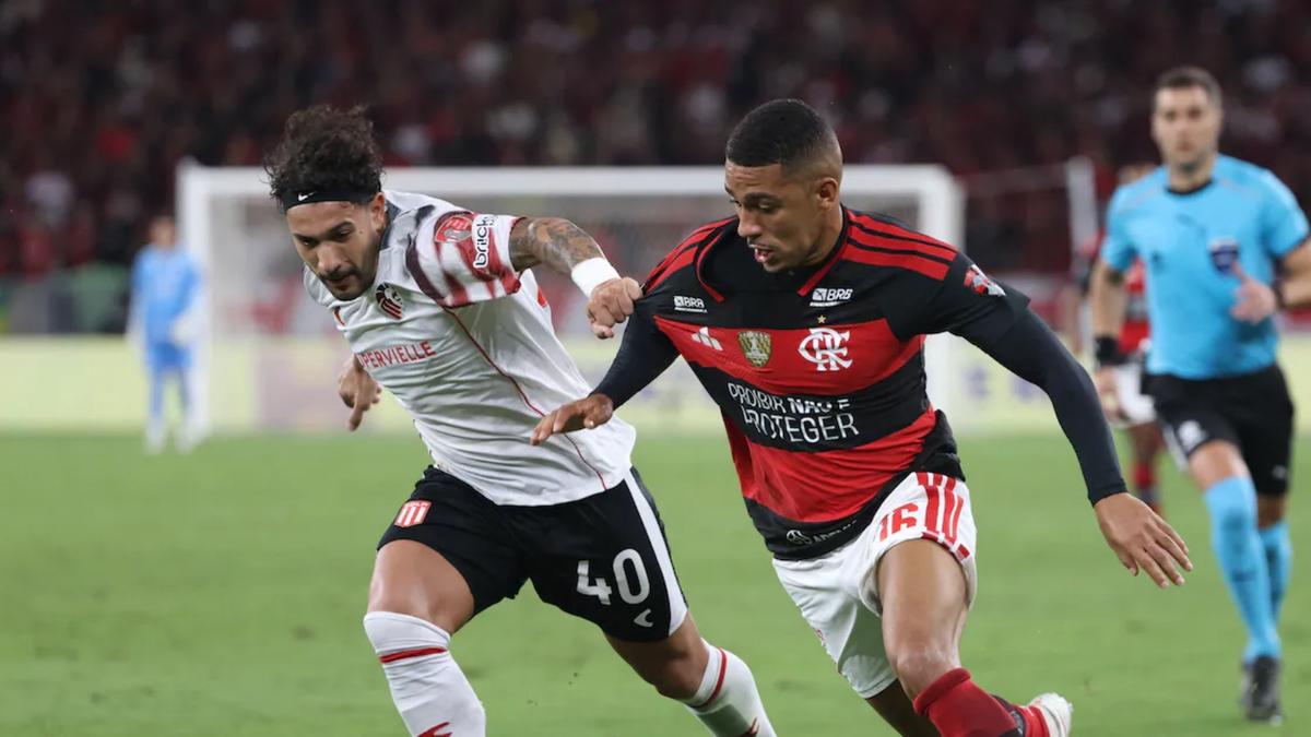 Dois jogadores disputam a bola em uma partida de futebol, com o árbitro ao fundo.