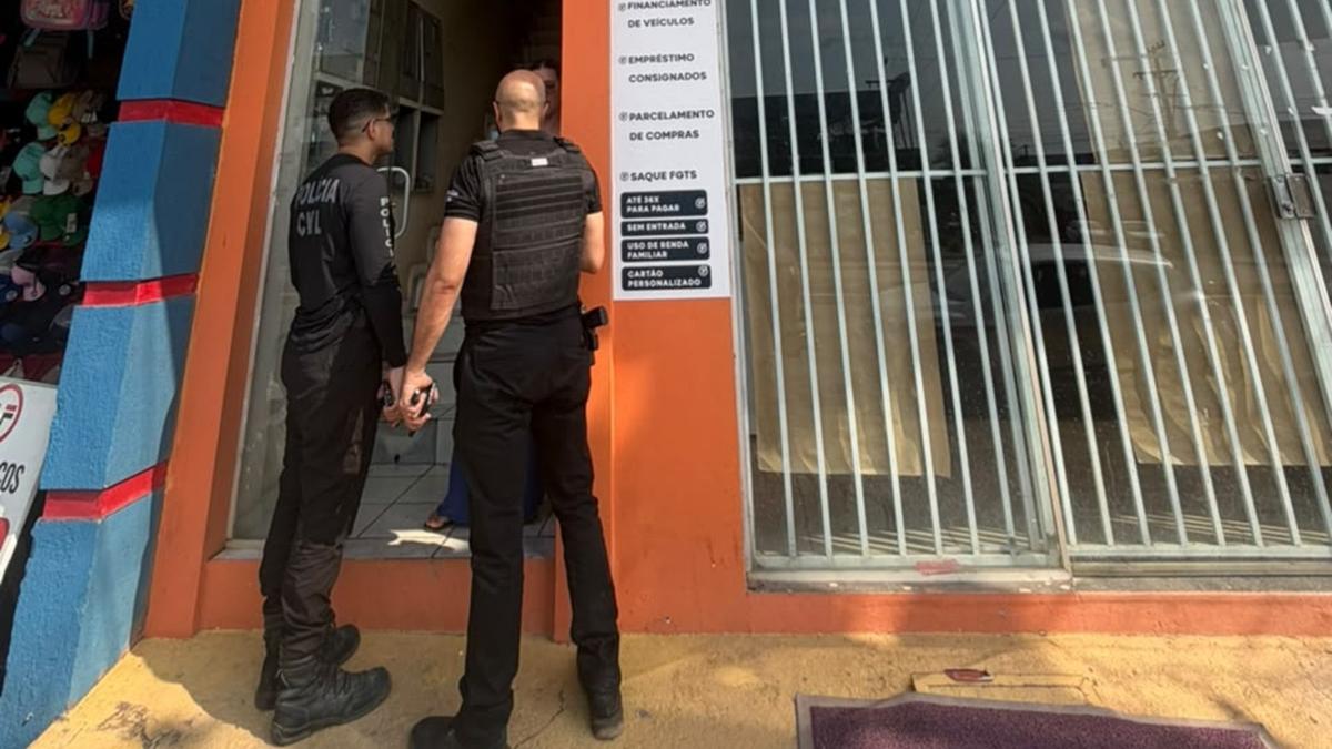 Dois policiais civis na entrada de um prédio com fachada laranja e placas comerciais.